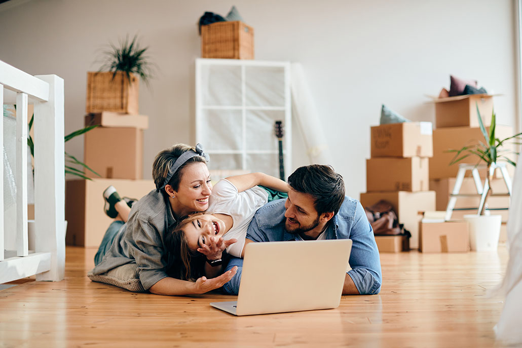 Happy parents and daughter having fun while using laptop and lying down on the floor after moving into new house.