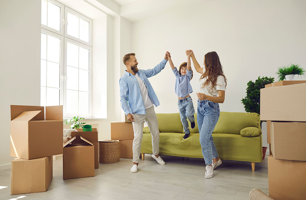 Happy family playing and having fun in new home full of cardboard boxes. Cheerful mom and dad holding son by the hands as he jumps off sofa in living room. Real estate, buying and moving house concept